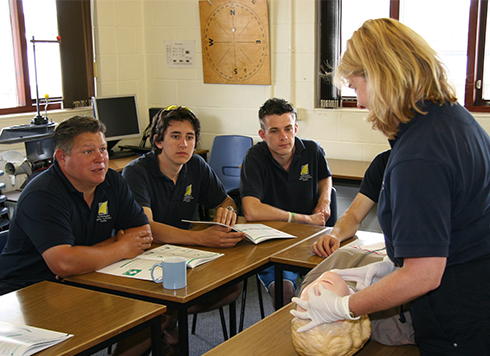 Men sat at desks listening to female instructor on RYA First Aid course